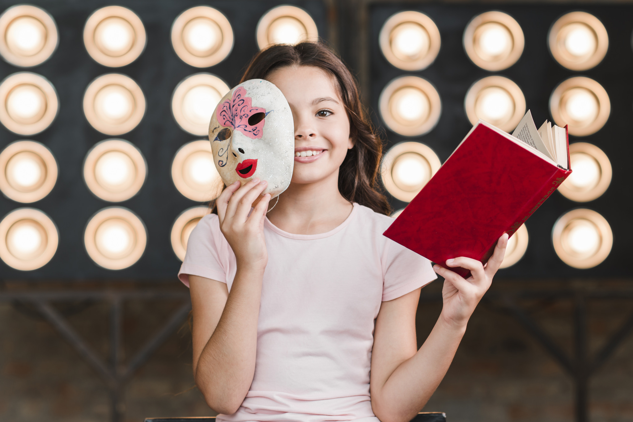 smiling-girl-sitting-front-stage-light-holding-mask-book